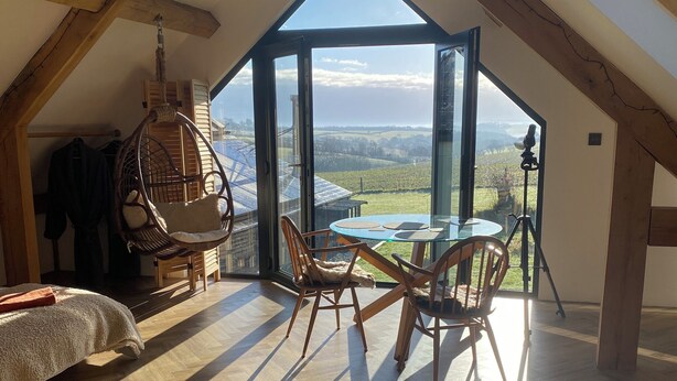 A spacious attic room with a large skylight, wooden beams, a hanging chair, a glass table, and a view of the countryside. - Exclusive Couples retreat surrounded by beautiful North Devon countryside. (Burrington)