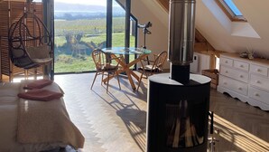 A cozy attic room with a large skylight, a wood-burning stove, a dining table with chairs, and a view of the countryside. - Exclusive Couples retreat surrounded by beautiful North Devon countryside. (Burrington)