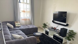 Una sala de estar moderna con un sofá seccional gris, una alfombra negra, un televisor de pantalla plana, una mesa de centro blanca y dos plantas en macetas.