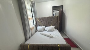 Una cama bien hecha con sábanas y almohadas blancas, un cabecero gris y una alfombra roja que conduce a una puerta.