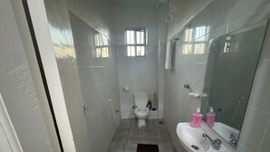 Un baño estrecho con inodoro, lavamanos y espejo. Hay una ventana sobre el inodoro y una zona de ducha a la derecha.