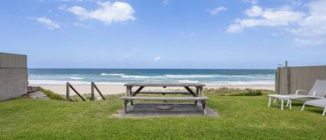 A grassy area with a picnic table and white lounge chairs facing a beach.