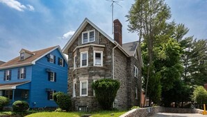 Une maison en pierre à deux étages, dotée d’une cheminée en pierre, une maison bleue attenante, et une pelouse bien entretenue avec un muret en pierre et un petit arbuste.