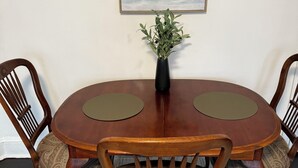 Une table de salle à manger ronde en bois, accompagnée de deux chaises, un vase rempli de feuillage vert et, au mur, un tableau représentant un paysage encadré.