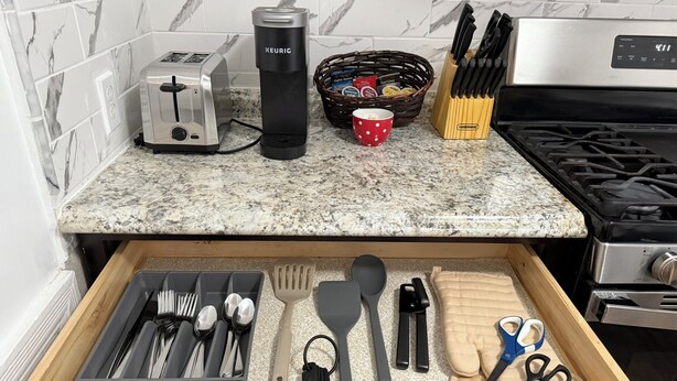 Un plan de travail de cuisine avec un grille-pain, une machine à café Keurig, un panier de sachets de thé, une tasse rouge et un bloc à couteaux.