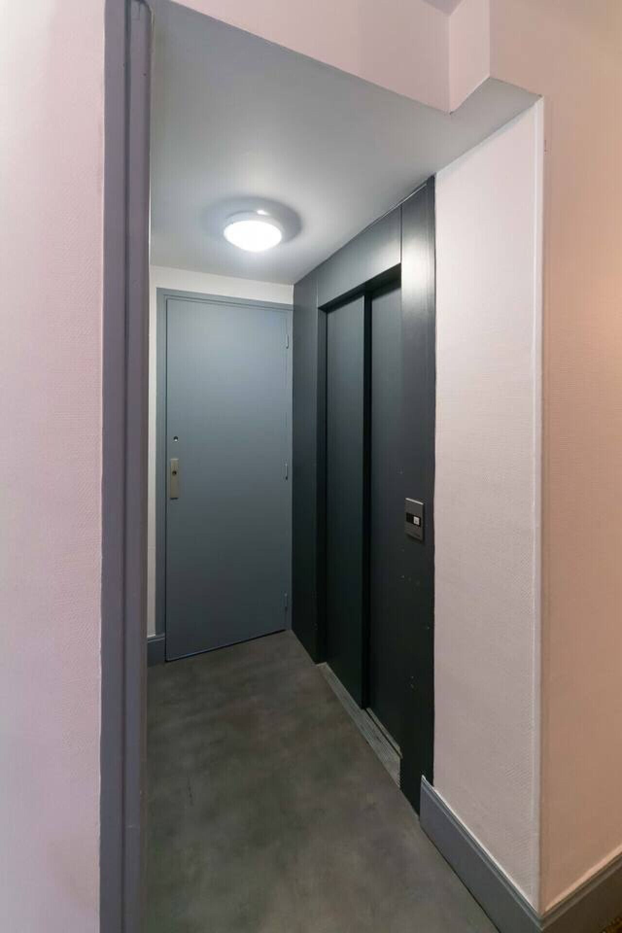 Un pasillo estrecho con alfombra gris, una puerta al final y una lámpara en el techo.