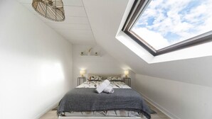 Ein Schlafzimmer mit Dachfenster, einem Bett mit grauer Decke, zwei Nachttischen mit Lampen und einer kleinen Ablage mit Dekoration.
