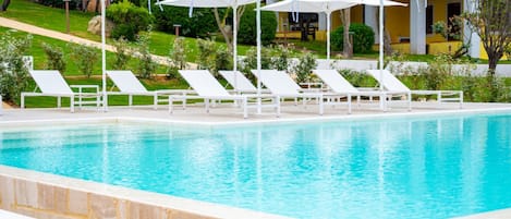 A swimming pool with white lounge chairs and umbrellas, surrounded by greenery and a building in the background.