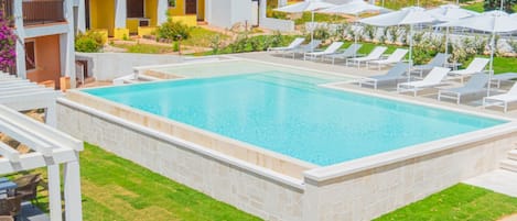 Ein Schwimmbecken mit Liegestühlen und Sonnenschirmen, umgeben von einer gepflegten Rasenfläche und einem mehrstöckigen Gebäude mit Balkonen.