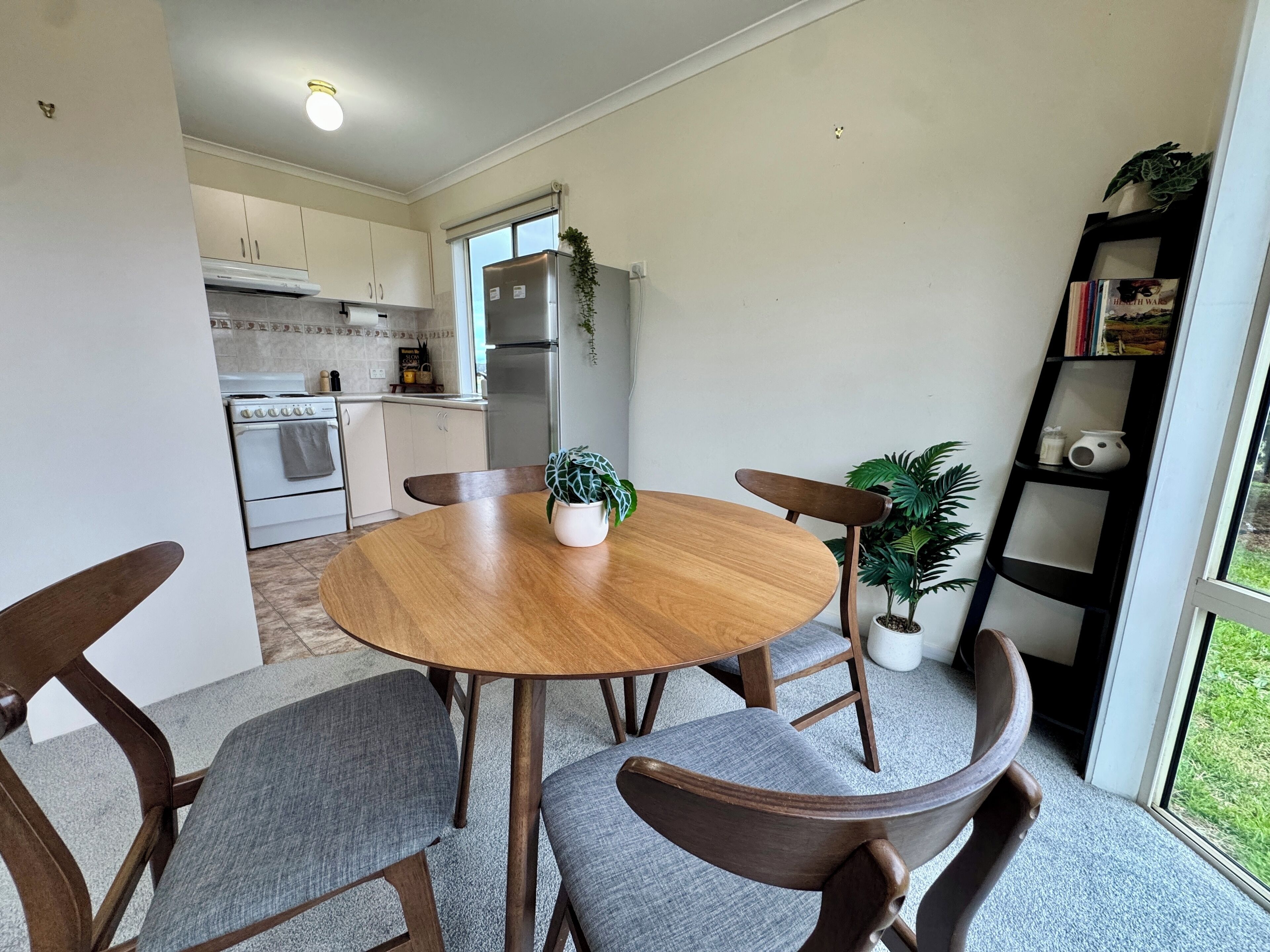 Un coin repas avec une table en bois et quatre chaises, une cuisine en arrière-plan et une bibliothèque contenant des livres et une plante.