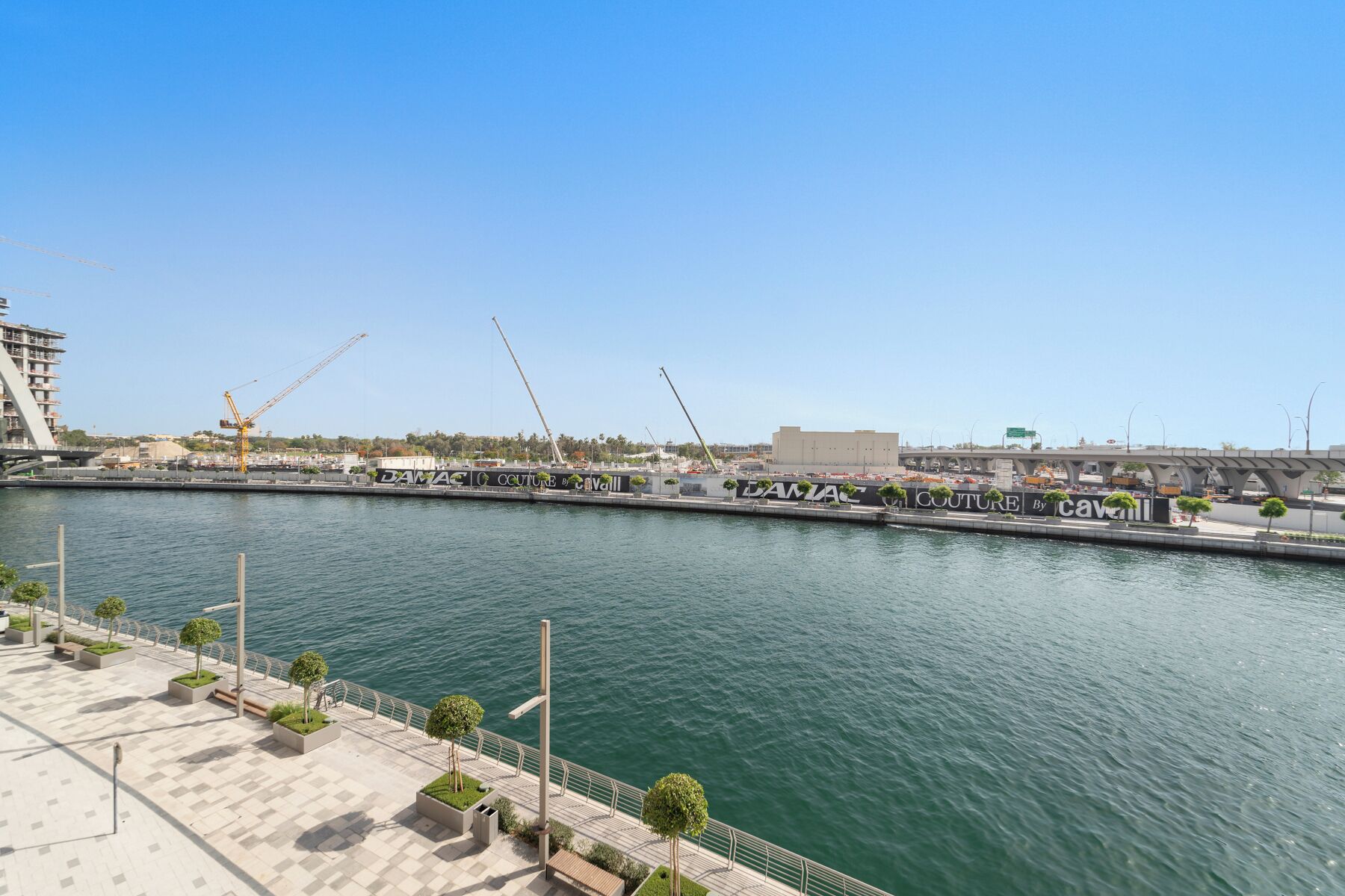 Eine Uferpromenade mit einer modernen Brücke, einem Yachthafen und Baukränen im Hintergrund.