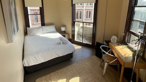 A bedroom with a bed, a desk, a chair, a ceiling fan, and large windows with a view of buildings. - GREAT VALUE COMFORT WITH THE PERFECT LOCATION! (Redfern)