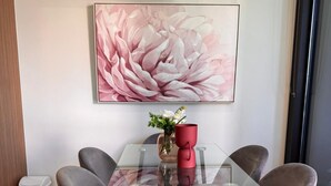 Uma área de jantar moderna com mesa de vidro cercada por quatro cadeiras cinzentas, um vaso vermelho com flores e um quadro grande com motivos florais rosa na parede.