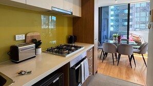 Cozinha moderna com fogão embutido, micro-ondas e torradeira. É visível uma área de refeições com mesa e cadeiras através de um painel de vidro.