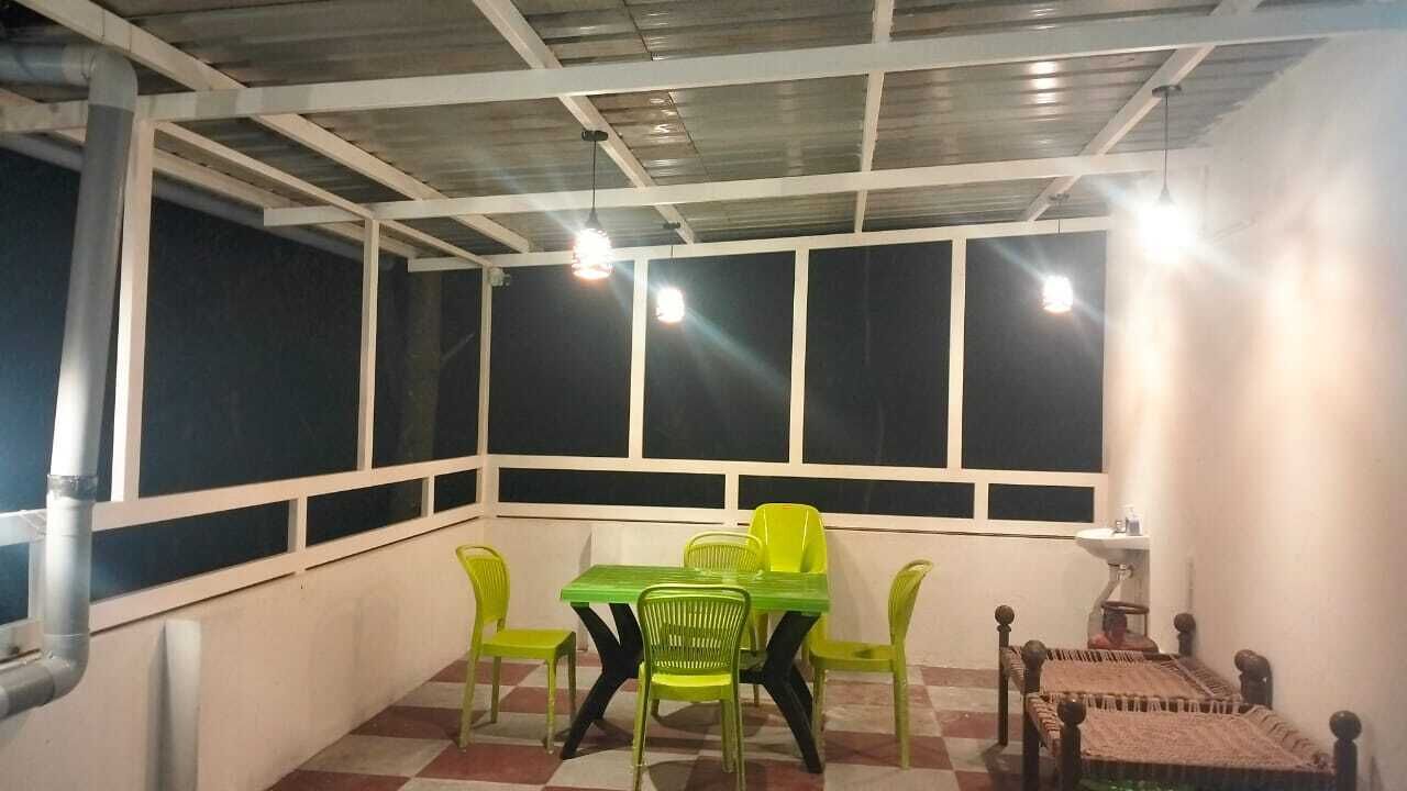 A room with a checkered floor, a green table, and four green chairs. There are three windows with white frames, and a bed with a patterned blanket is visible.