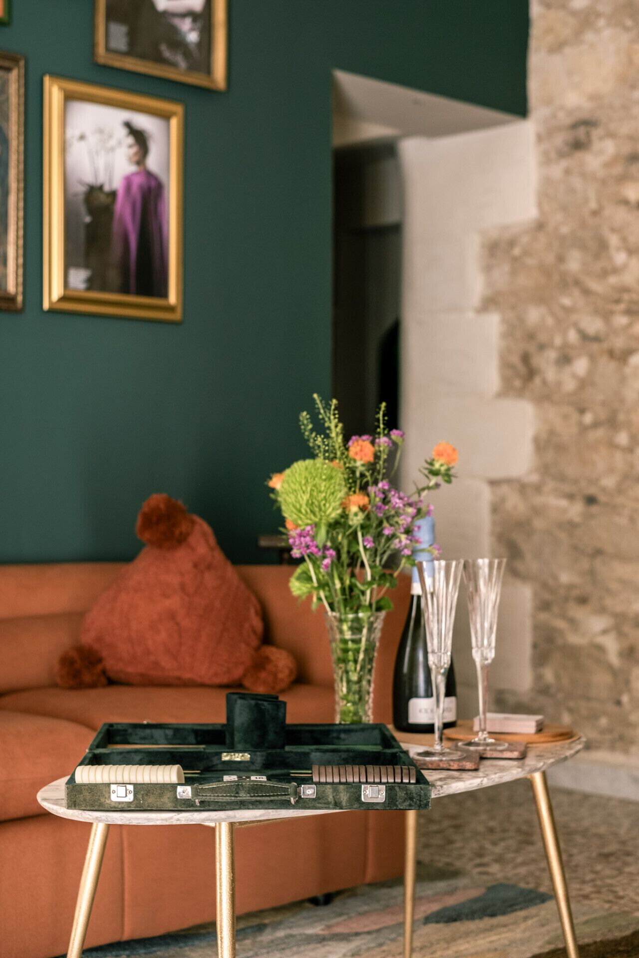 Een gezellige woonkamer met een groene wand, een bruine bank, een kleine tafel met een bloevas, en een fles wijn.