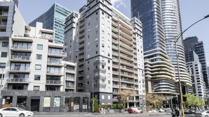 Moderni kaupunkikatu, jossa on korkeita kerrostaloja, mukaan lukien kaareva lasinen pilvenpiirtäjä ja useita kerroksia sisältävä asuintalo, jossa on parvekkeita.