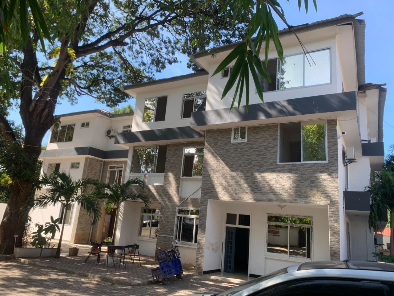 Un edificio moderno de varios pisos con techo plano, ventanas amplias y un acceso pavimentado con algunas plantas en macetas.