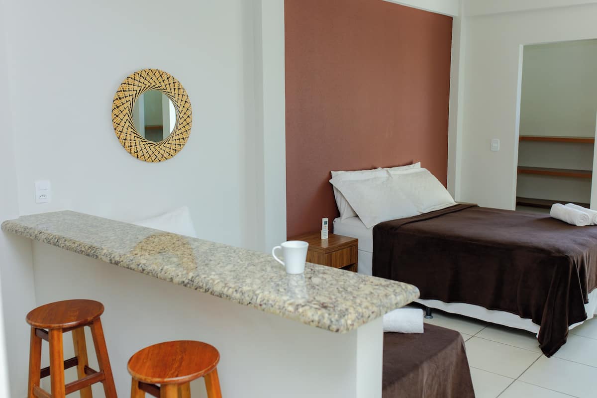 Camera d'hotel con un piano di lavoro in granito, due sgabelli in legno, un letto con coperta marrone, un cuscino bianco e uno specchio rotondo con motivo geometrico.