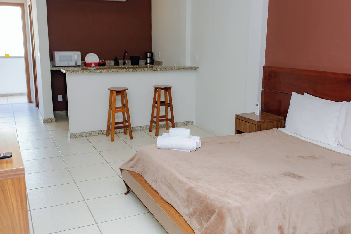 Una camera d'hotel con un letto, un comodino in legno, uno sgabello di legno e un angolo cottura con un forno a microonde e un lavandino.