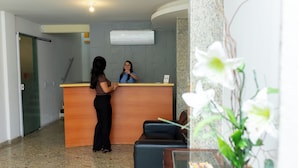 A reception area with a wooden counter, a receptionist, and a guest standing. - Pousada Caiçara Flats (Cabo Frio)