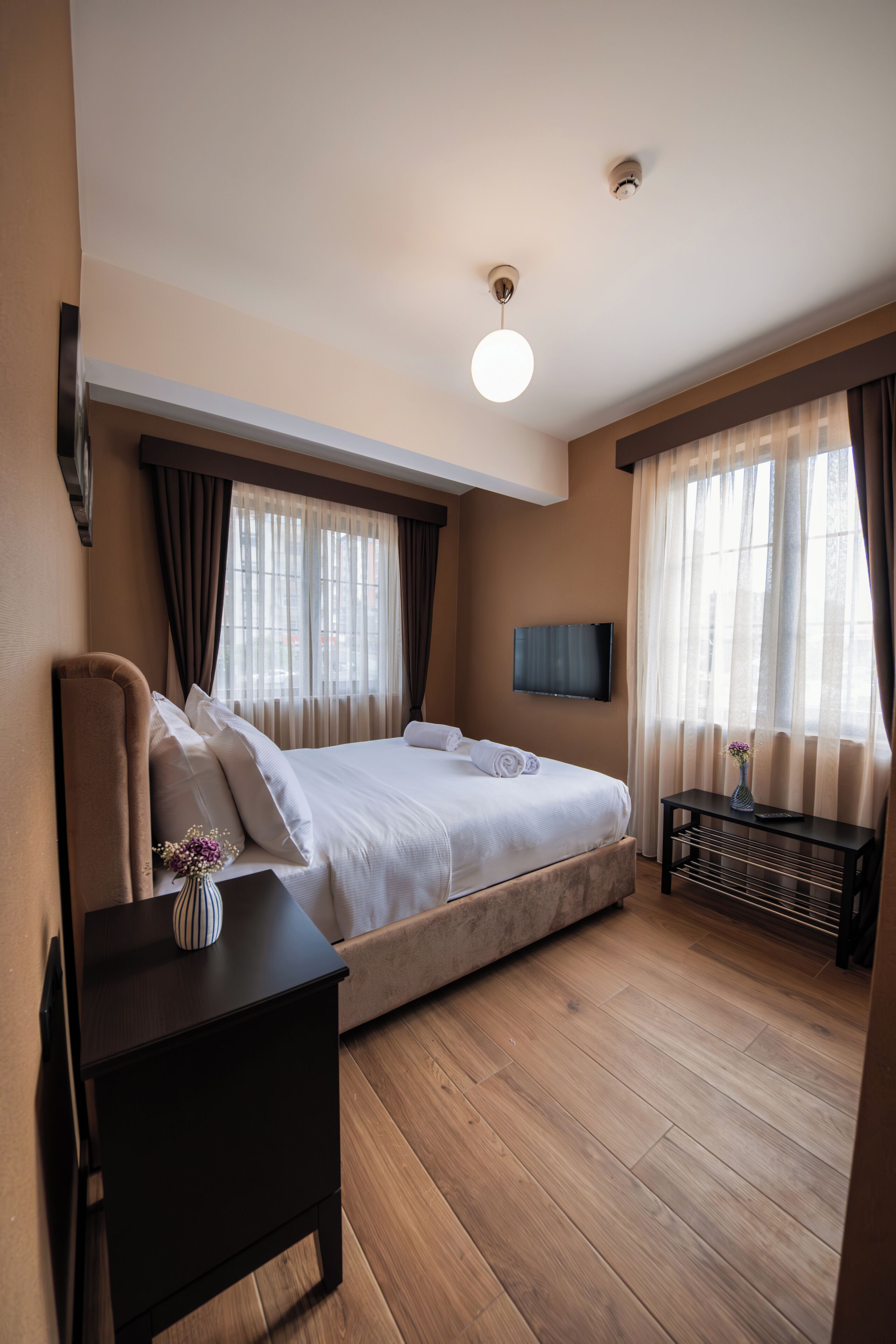 Una habitación de hotel con cama, mesita de noche, televisión montada en la pared y ventana con cortinas.