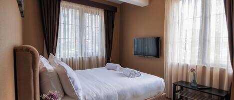 Una habitación de hotel con cama, mesita de noche, televisión montada en la pared y ventana con cortinas.