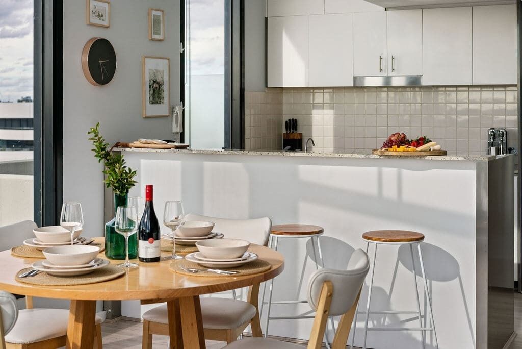 Une cuisine moderne avec des meubles blancs, un coin repas avec une table en bois et des chaises blanches, et une vue sur la ville grâce à de grandes fenêtres.