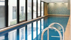 Una piscina interna con pareti in vetro trasparente, pavimento piastrellato e scaletta in metallo.