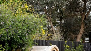 A wooden barrel structure with a door, surrounded by a garden with various plants and trees. - The Pavilion in Mount Martha (Melbourne)