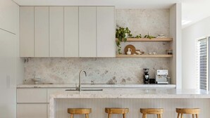 A modern kitchen with white cabinets, a marble countertop, wooden stools, and a coffee machine. - The Pavilion in Mount Martha (Melbourne)