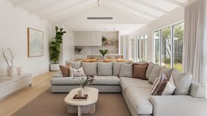 A modern living room with a large L-shaped sectional sofa, a marble coffee table, and a view of the kitchen and outdoors through large windows. - The Pavilion in Mount Martha (Melbourne)
