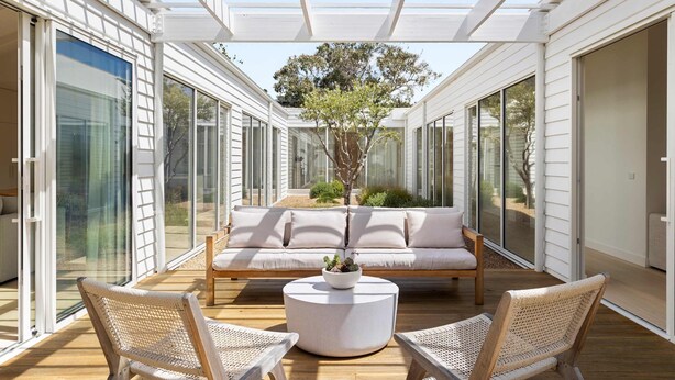 A sunlit patio with a sofa, two chairs, and a round table, surrounded by glass doors and a tree. - The Pavilion in Mount Martha (Melbourne)