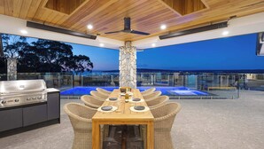 An outdoor dining area with a stone pillar, a grill, wicker chairs, and a view of the sea at dusk. - Baymere Pool Spa City Views (Melbourne)