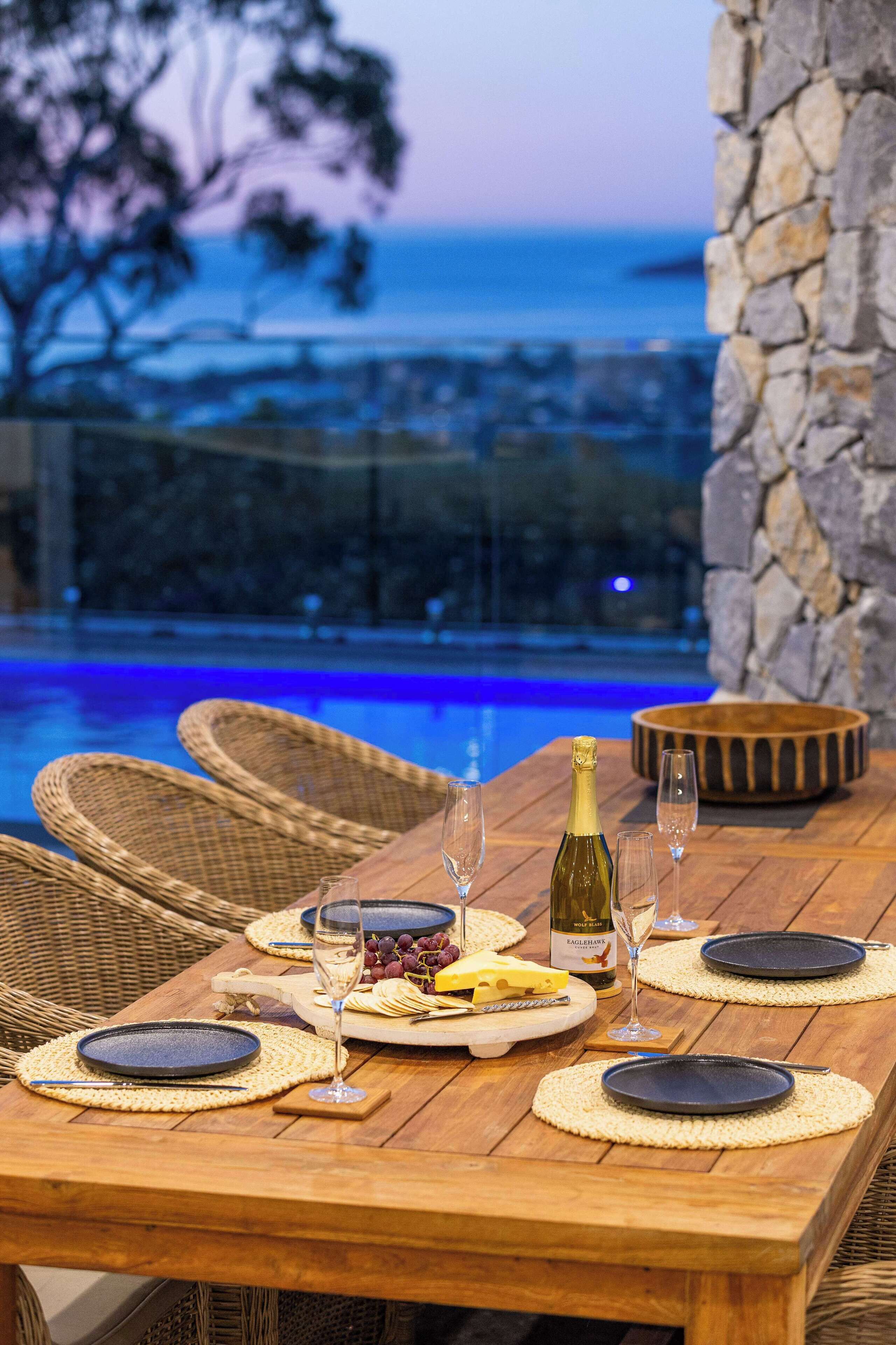 A wooden dining table set for a meal with a bottle of wine, glasses, and plates, overlooking a pool and ocean view.