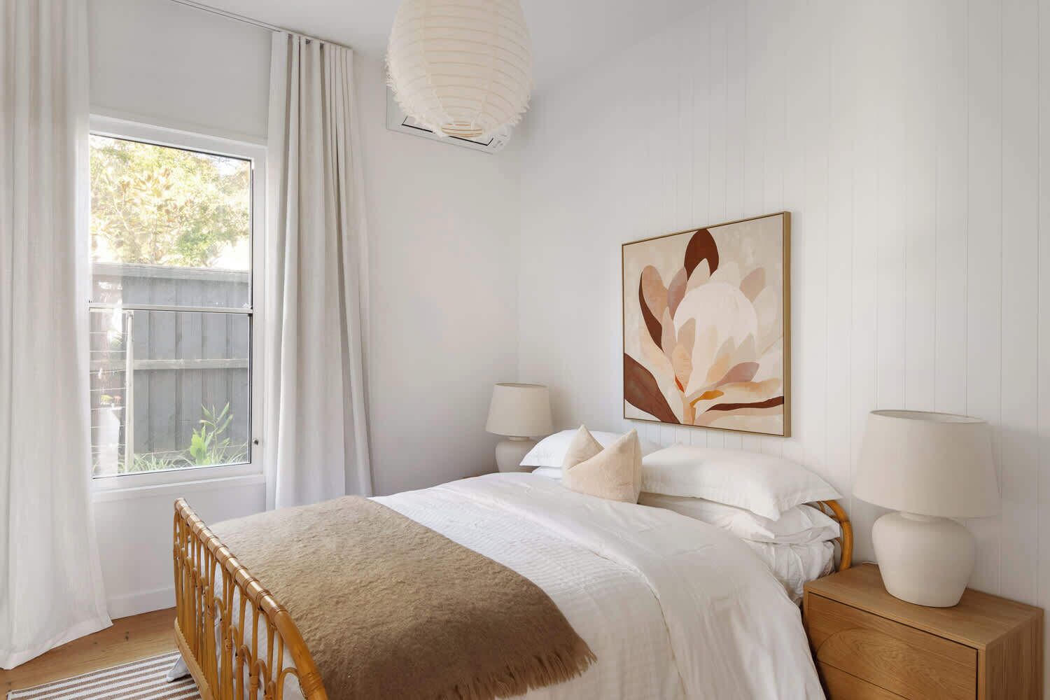 A bedroom with a bed, two bedside lamps, a large abstract painting, and a window with curtains.