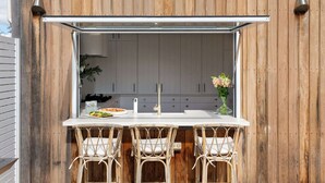 Una cocina al aire libre con encimera blanca, tres sillas de mimbre y una mesa de comedor.