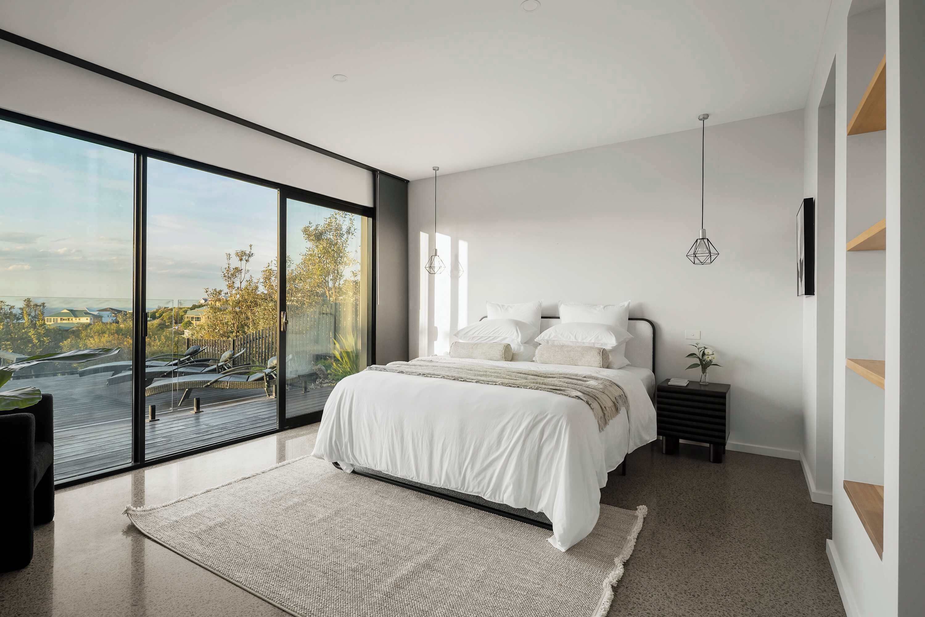 A modern bedroom with a large sliding glass door leading to a balcony, a bed with white bedding, a small bedside table, and a hanging light fixture.