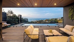 Et moderne udendørs loungeområde med træterrasse, polstrede siddepladser og udsigt til en swimmingpool og det fjerne landskab.