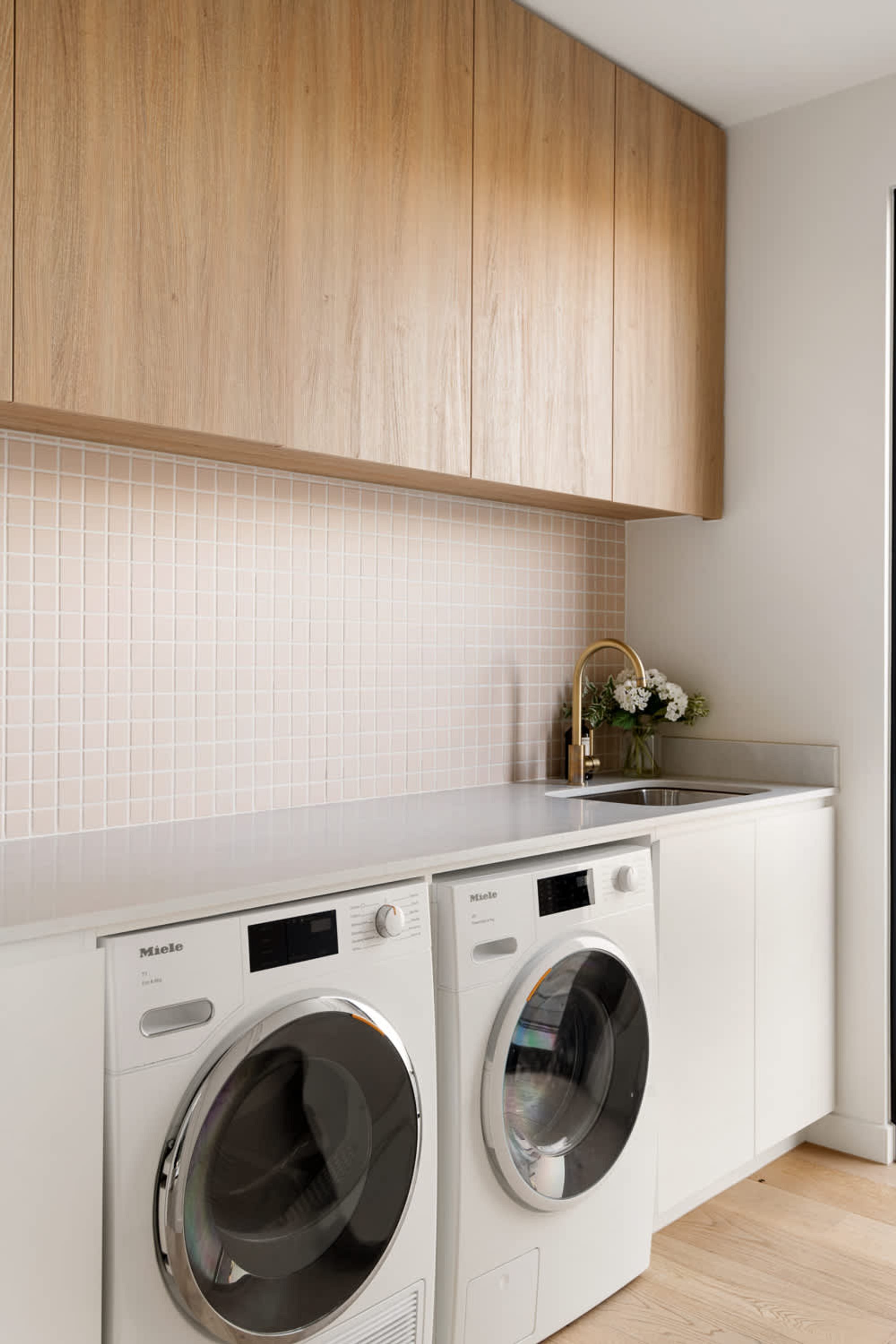 A modern kitchen with wooden cabinets, a white countertop, a washing machine, and a sink with a vase of flowers.