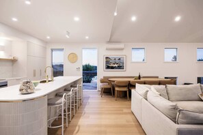 Una cocina moderna con una isla central, taburetes de bar, un comedor con una mesa y sillas de madera, y una sala de estar con un sofá.