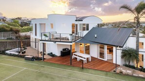 Une maison moderne à deux étages, dotée d’un balcon, d’une terrasse avec mobilier d’extérieur et d’un court de tennis.