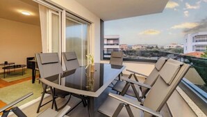 Un balcon avec une table et des chaises en verre, une petite table avec un vase, et une vue sur des bâtiments au loin.