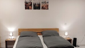 A bed with a wooden frame, two pillows, a grey blanket, and a brown rug. There are two bedside tables with lamps, and three framed pictures on the wall. - Holly-WOOD Allgäu vacation apartment - relaxation with heart in the countryside (Markt Rettenbach)