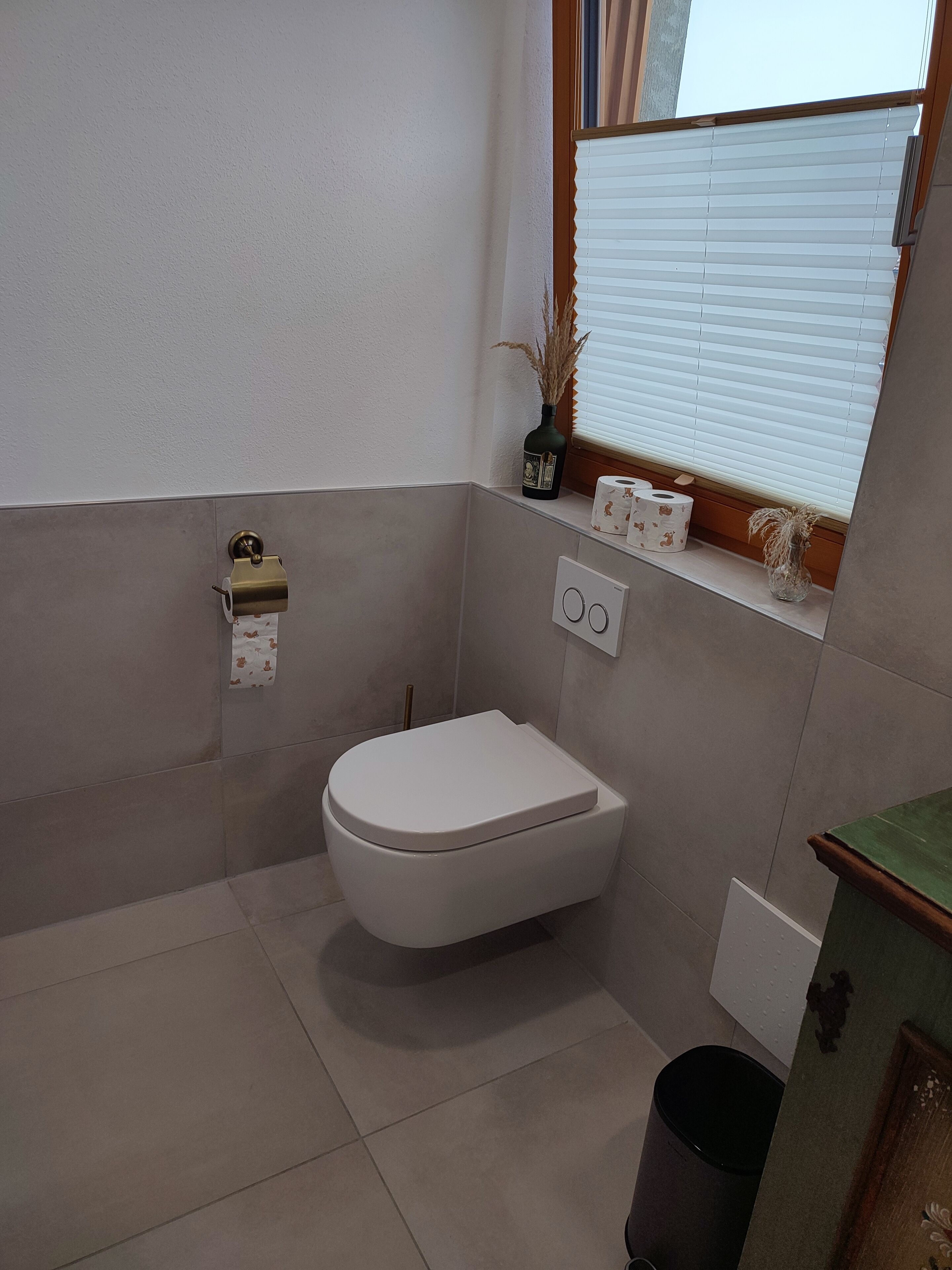 A compact bathroom with a white toilet, a small window with blinds, a tissue dispenser, and a trash bin.