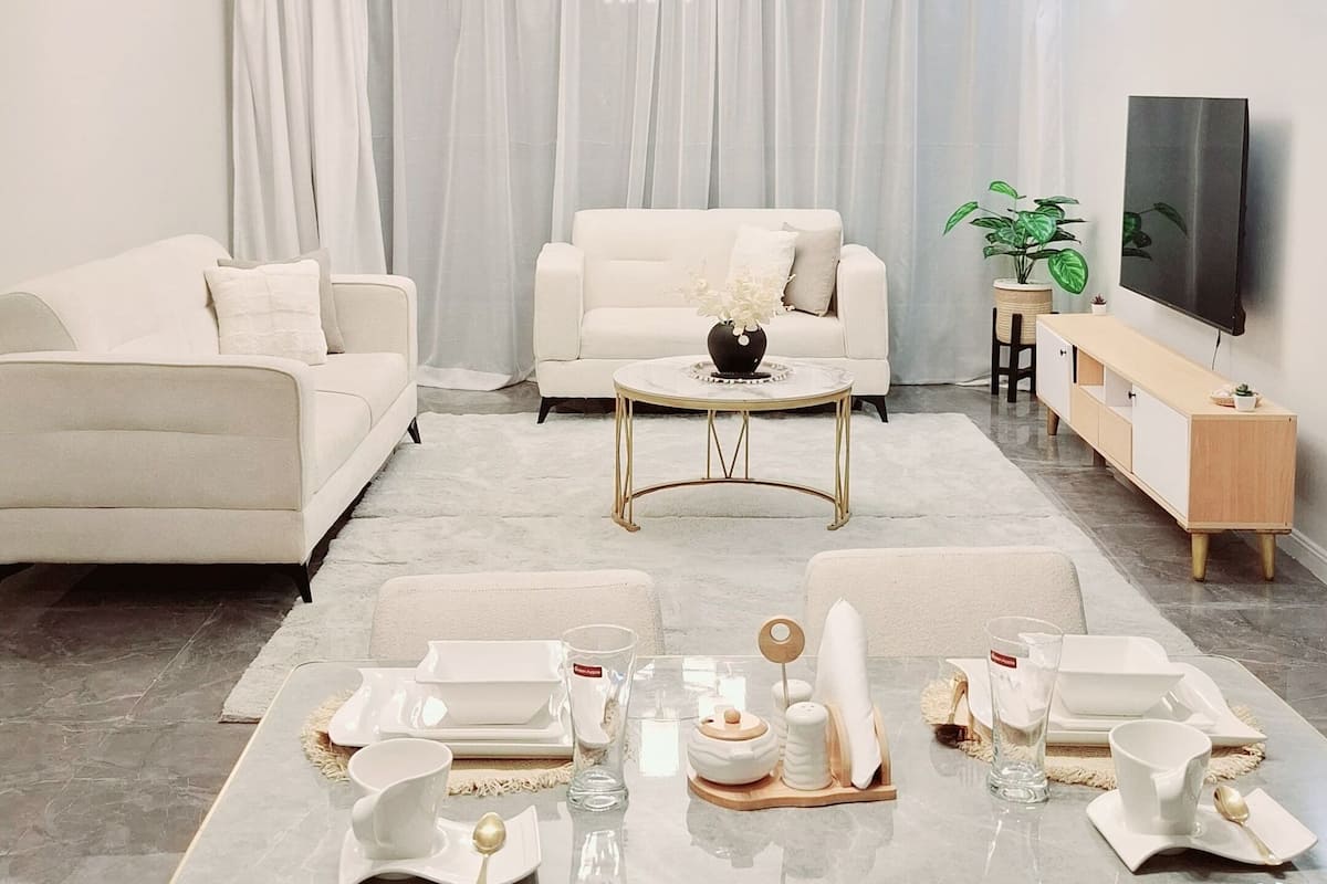 Ein modernes Wohnzimmer mit Essbereich, ausgestattet mit einer Sofagarnitur, einem Kronleuchter, einem Flachbildfernseher und einem Esstisch mit Tellern und Gläsern.