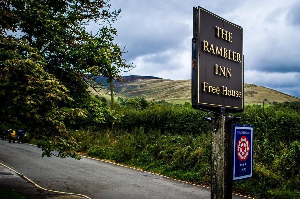 Letrero de The Ramblers Inn, pub tradicional, con una vista panorámica de colinas y una carretera, con gente al fondo.