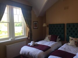 Una habitación de hotel con dos camas, una ventana con cortinas, un radiador, una lámpara fijada a la pared y un cuadro enmarcado.