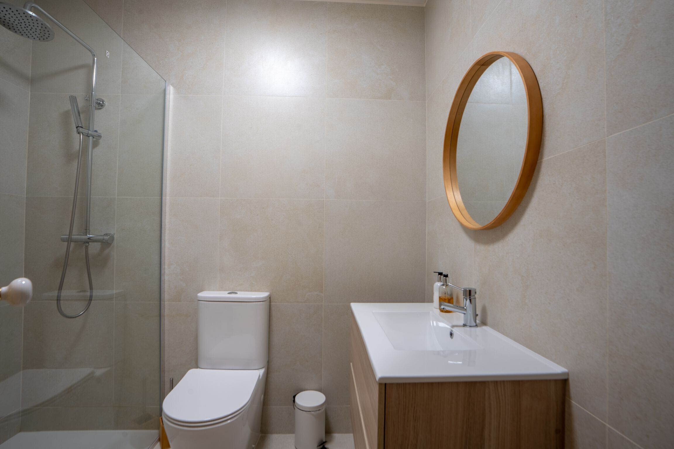 Ein modernes Badezimmer mit einer gläsernen Duschkabine, einer weißen Toilette, einem hölzernen Waschtisch mit Waschbecken und einem runden Spiegel an der Wand.