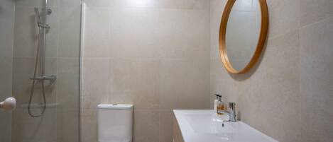 Ein modernes Badezimmer mit einer gläsernen Duschkabine, einer weißen Toilette, einem hölzernen Waschtisch mit Waschbecken und einem runden Spiegel an der Wand.